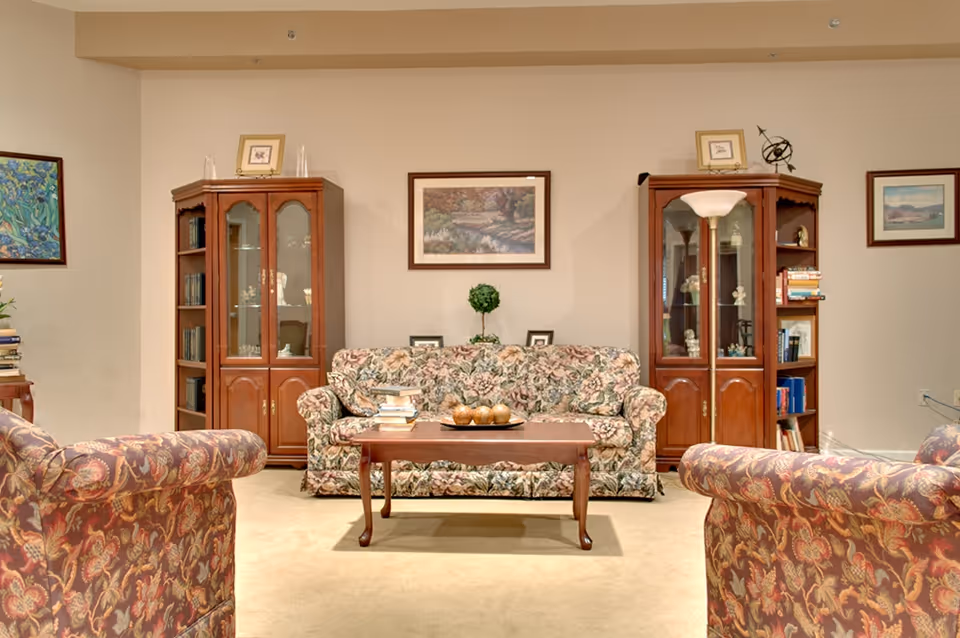 A cozy living room with floral patterned sofas and armchairs arranged around a wooden coffee table. Behind the sofa, there are two wooden display cabinets filled with books and decorative items. The walls are adorned with framed paintings and a floor lamp stands next to one of the cabinets.