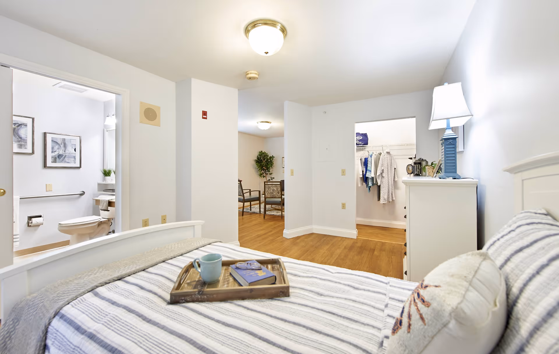 Bright furnished bedroom with a striped bed and tray, dresser with lamp, and open view into a bathroom and walk-in closet.
