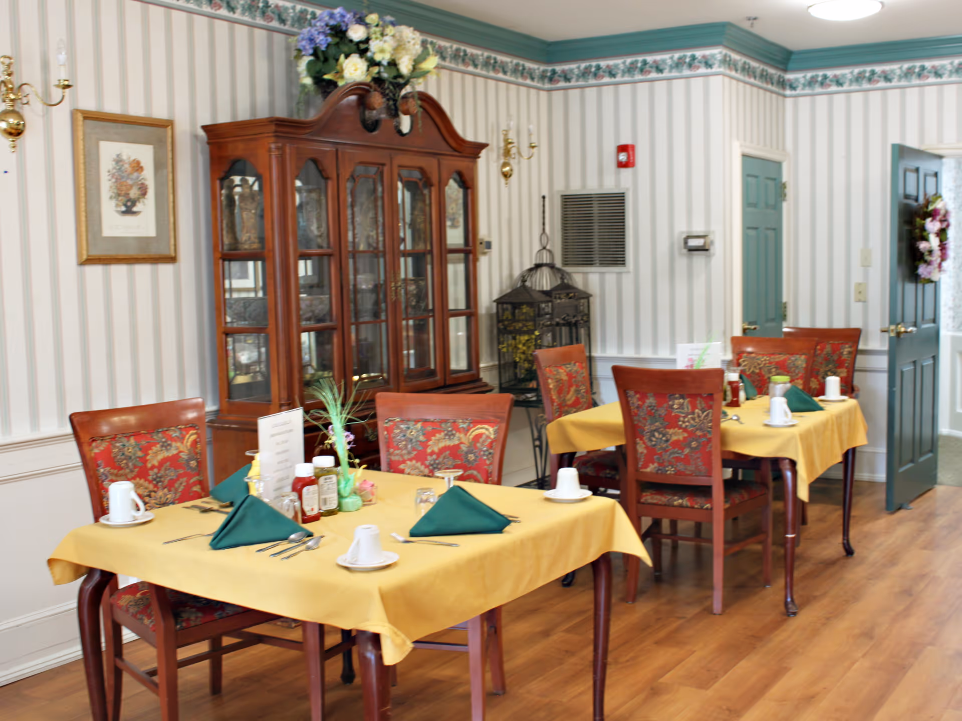 Dining room with tables set with yellow tablecloths and green napkins, floral-upholstered chairs, and a wooden china cabinet against striped wallpaper.