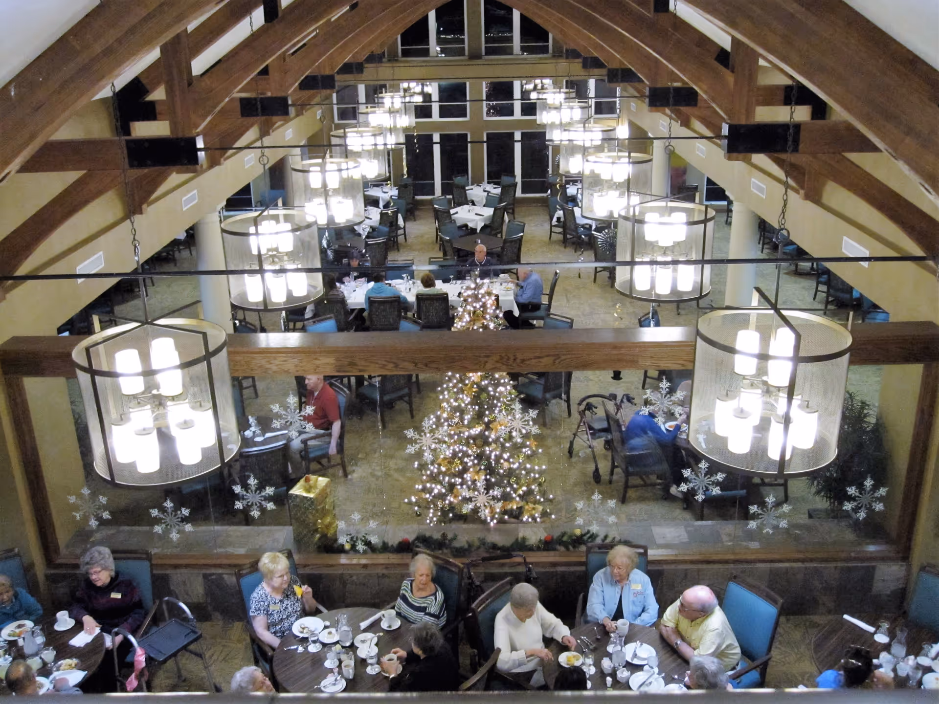 Interior view of a dining room in Stone Oak Retirement Community decorated for the holidays with a Christmas tree and snowflake decals on the glass partition. Several elderly people are seated at round tables enjoying a meal, with large hanging light fixtures and wooden beams visible on the ceiling.