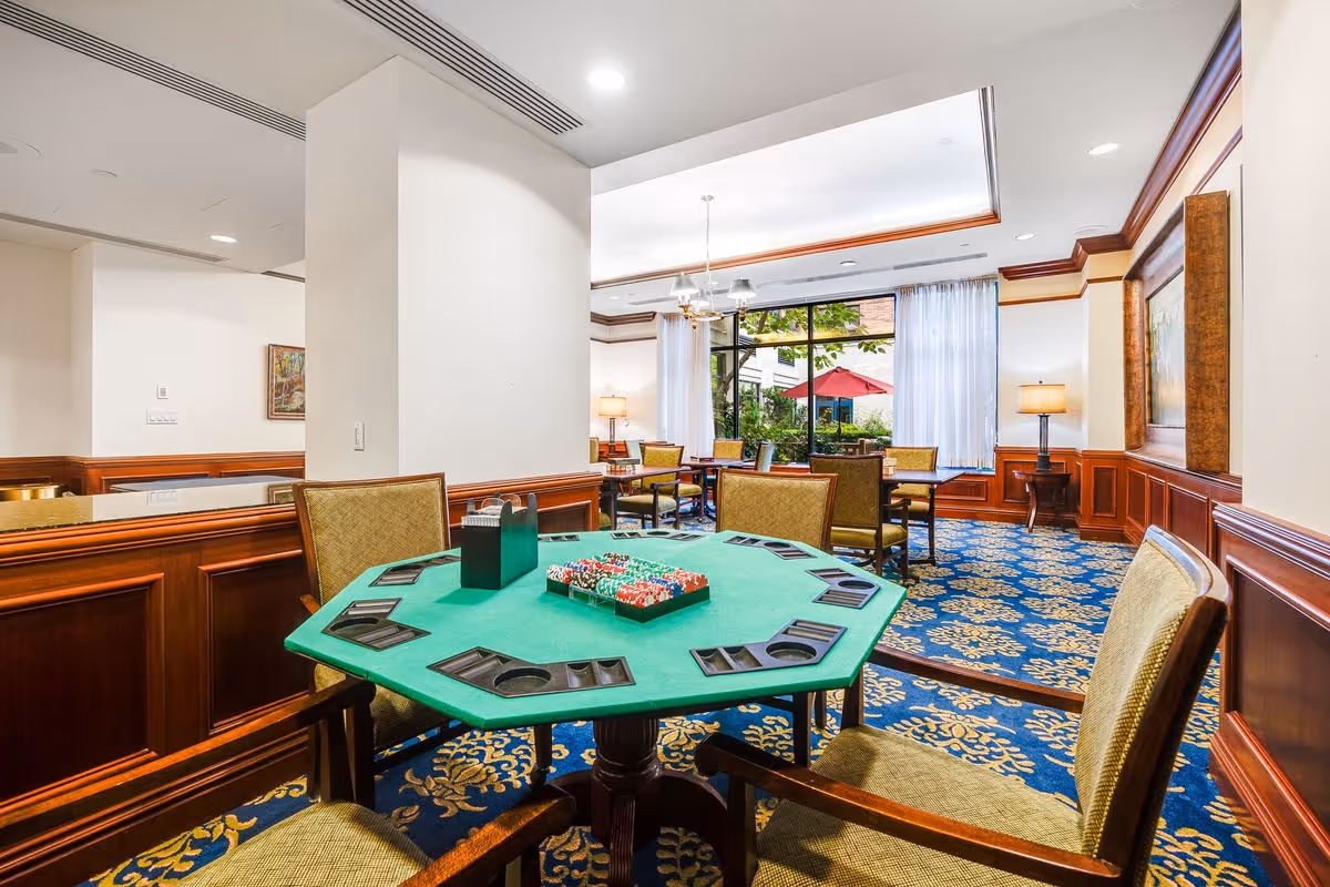 A well-lit interior room with a green poker table in the foreground surrounded by four upholstered chairs. The table has poker chips and card holders. In the background, there are additional tables and chairs near large windows with white curtains, allowing natural light to enter. The room features wood paneling, a blue carpet with a yellow floral pattern, and a ceiling light fixture.