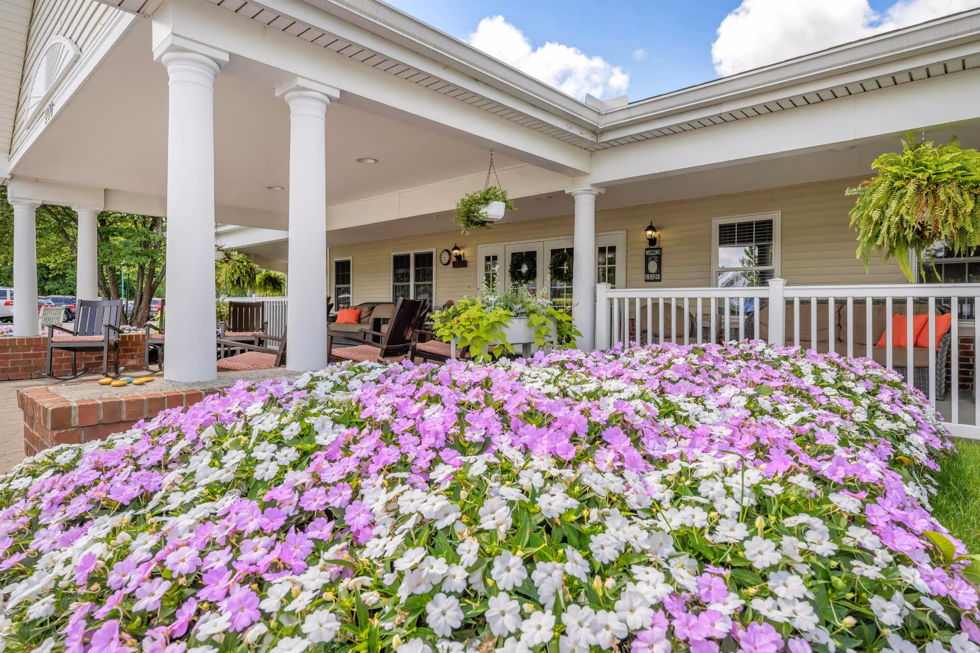 Outdoor patio area of a senior living facility with white columns, hanging plants, and a large flower bed filled with pink and white flowers in the foreground. There are chairs and seating areas under the covered porch with a welcoming atmosphere.