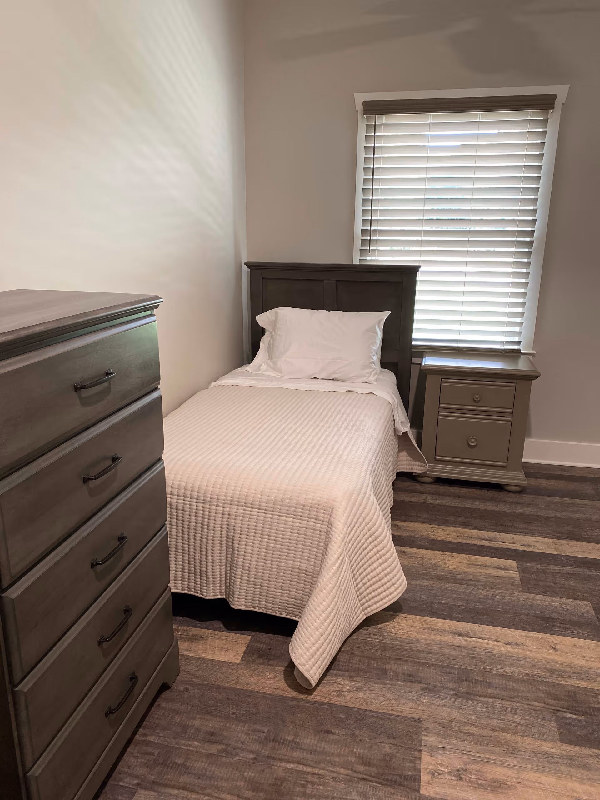 A small bedroom with a single bed covered in a white quilt and a white pillow. Next to the bed is a beige nightstand with two drawers. On the left side of the image is a tall dresser with five drawers. The room has wood flooring and a window with closed blinds behind the bed.