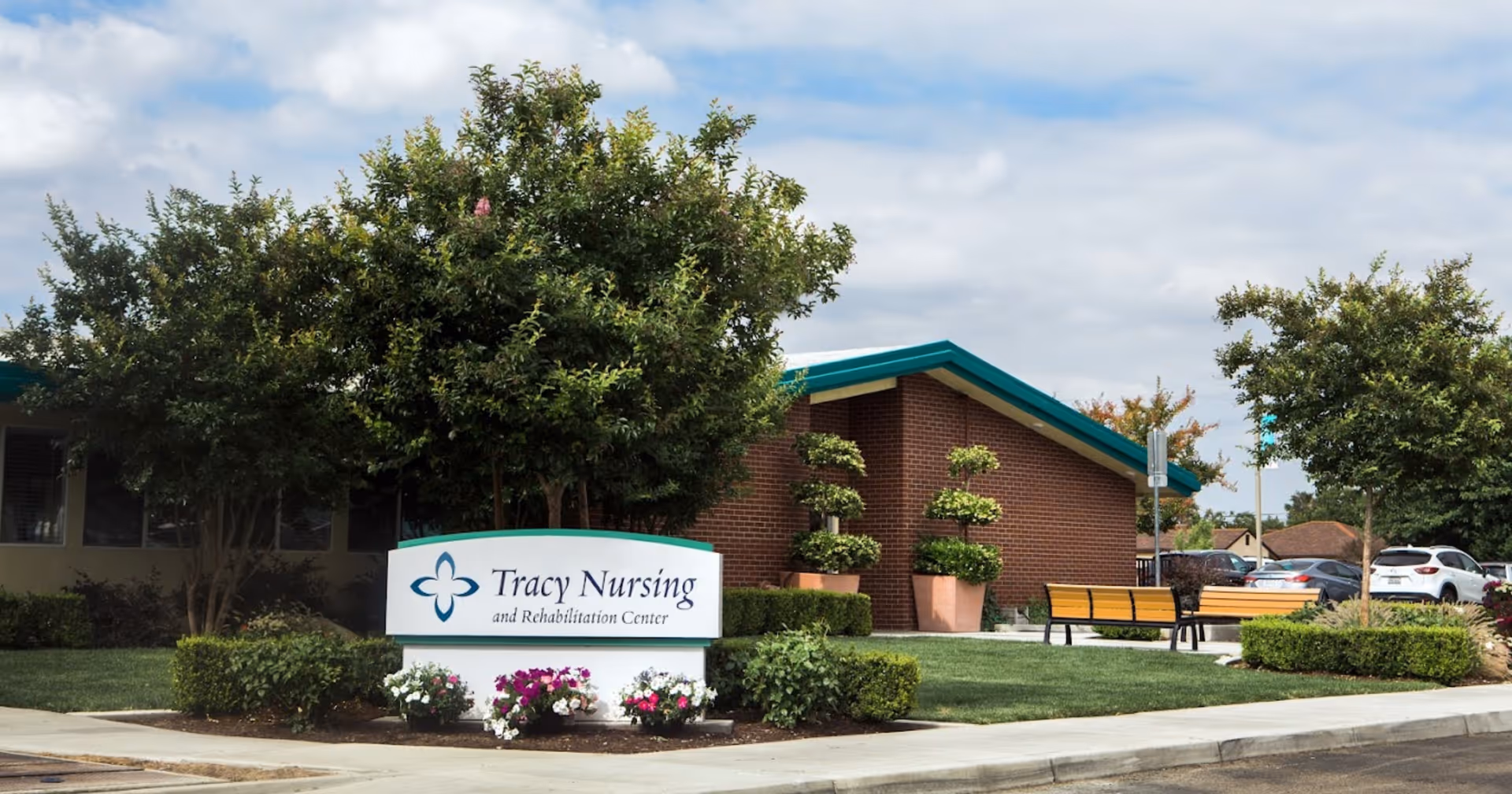 Front exterior of Tracy Nursing and Rehabilitation Center showing the facility sign, landscaped shrubs, benches, and a low brick building.