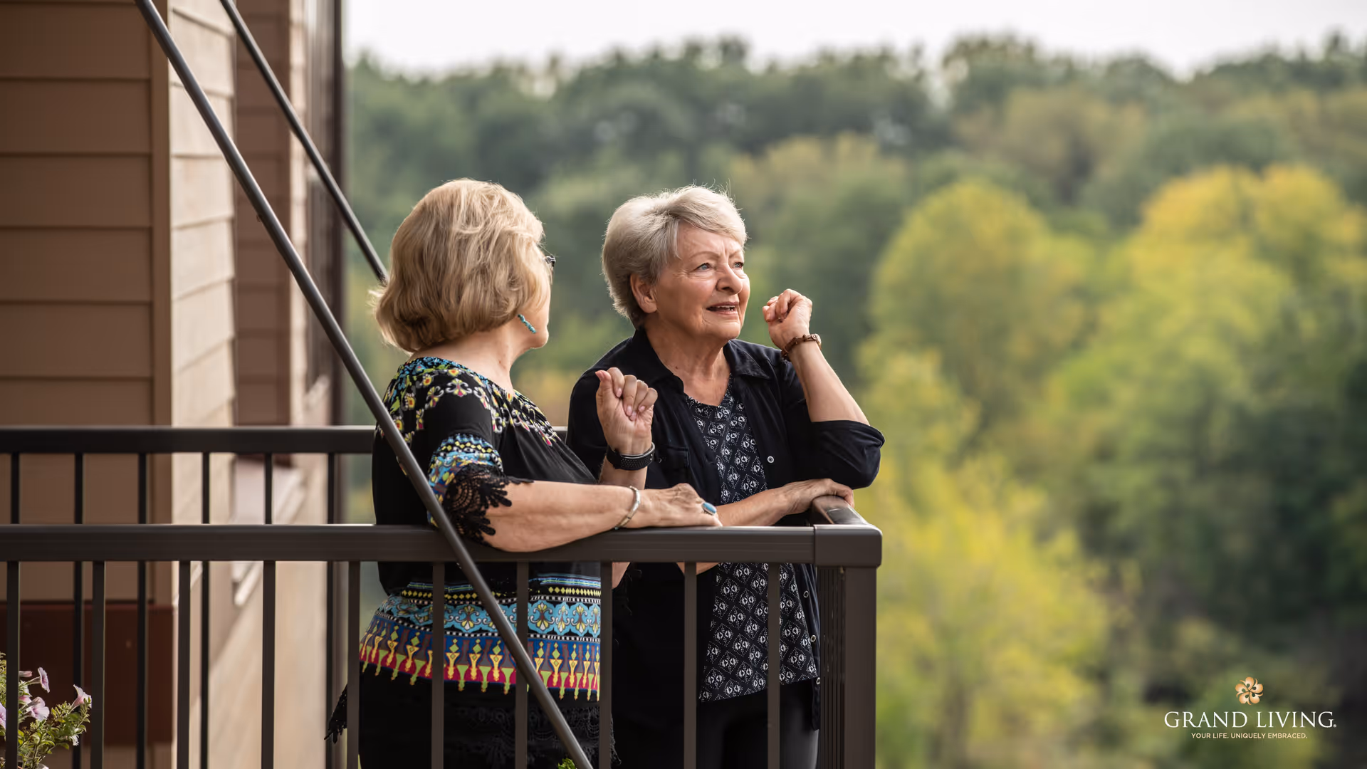 Two elderly women standing and talking on a balcony with a view of green trees in the background during daytime.
