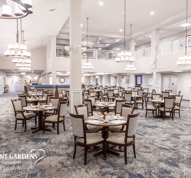 Large bright dining room with many round tables and chairs arranged under hanging chandeliers in a senior living facility.