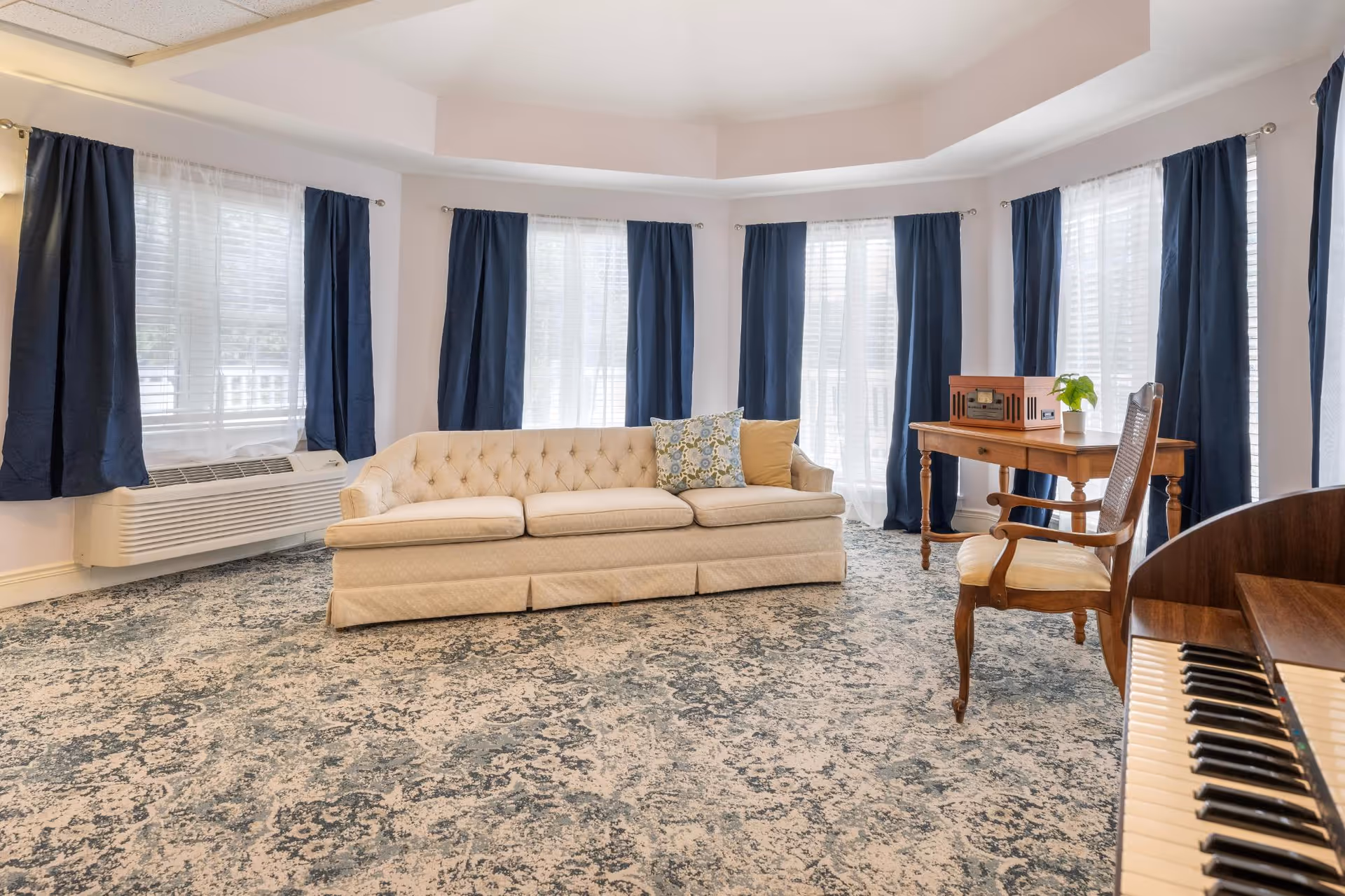Bright common living room with a tufted beige sofa, wooden desk and chair, piano keyboard at the edge, and tall windows with navy curtains.