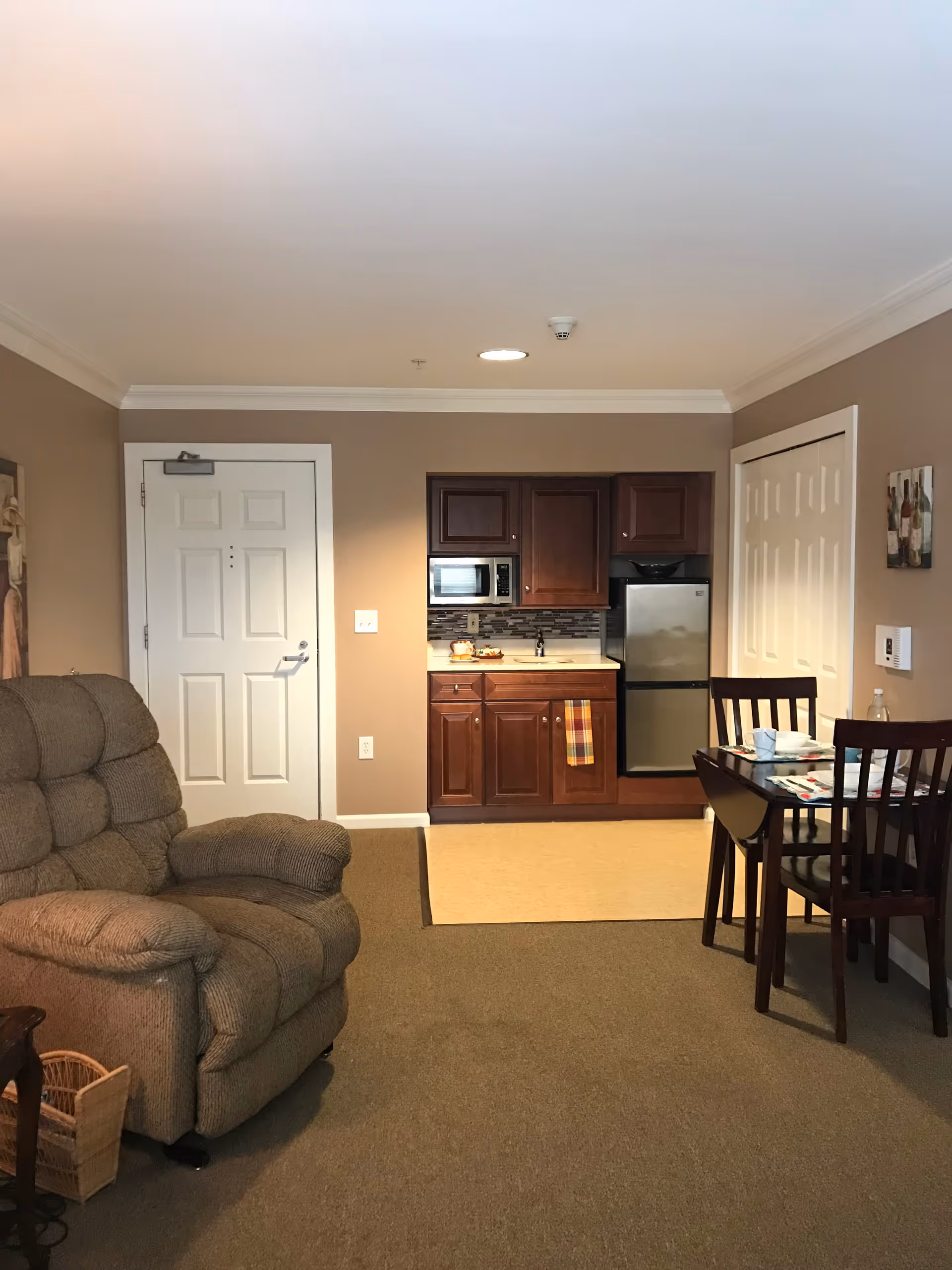 Interior view of a senior living facility room showing a small kitchen area with wooden cabinets, a microwave, a mini refrigerator, and a sink. To the right, there is a dining table set with two chairs and place settings. On the left side, there is a cushioned recliner chair. The walls are painted beige, and there is a white door and a closet with white double doors.