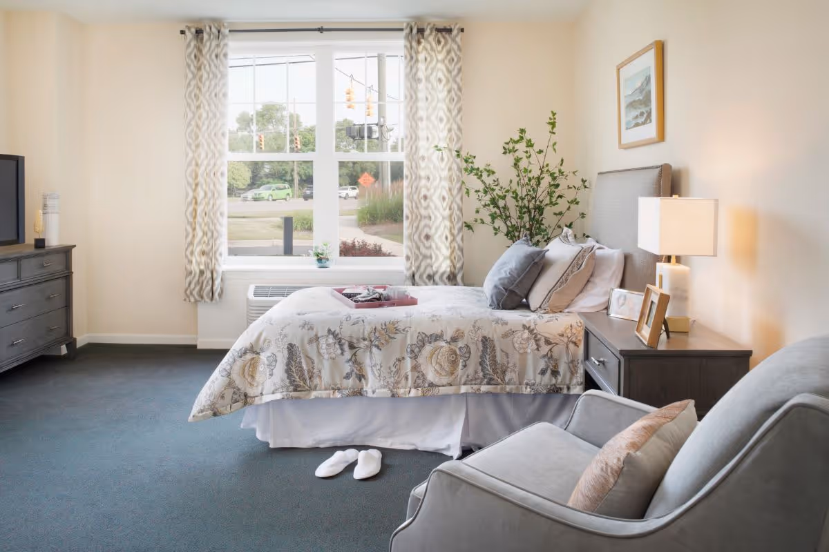 A cozy bedroom in a senior living facility with a bed covered in floral patterned bedding, a gray armchair with a pillow, a nightstand with a lamp and framed photos, a dresser with a TV, and a large window with patterned curtains letting in natural light. White slippers are placed on the carpeted floor beside the bed.