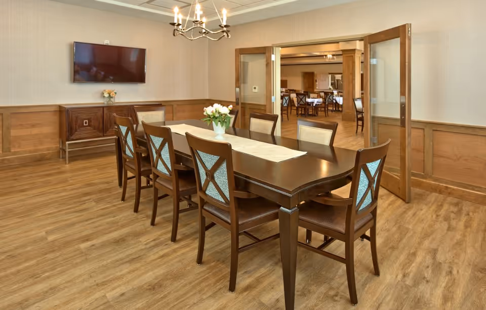 A long wooden dining table with eight chairs in a well-lit dining room with a wall-mounted TV and glass doors opening to another dining area.