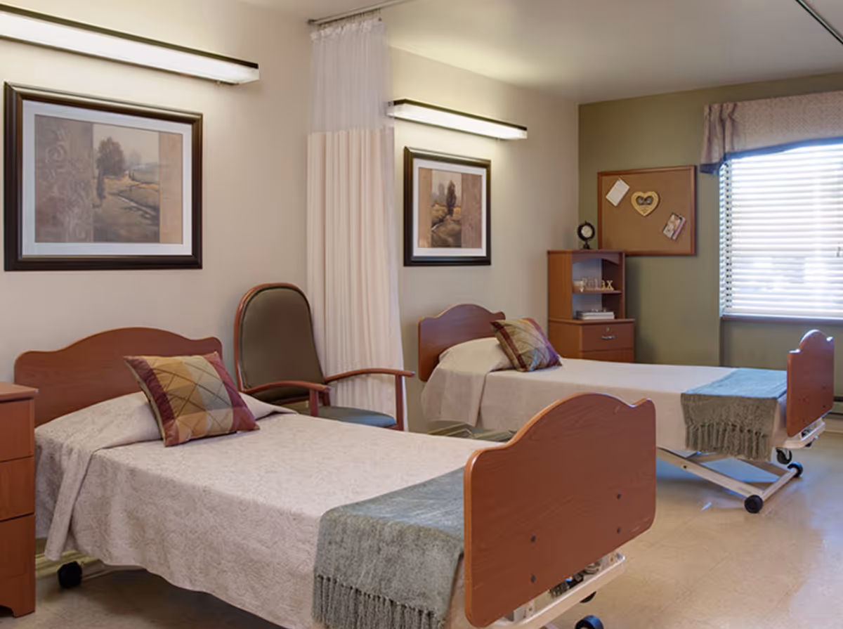 Two single hospital-style beds in a tidy shared resident room with bedside tables, chairs, framed artwork, and a window with blinds.
