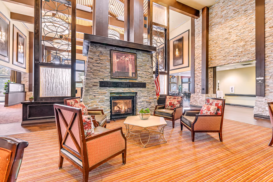 A cozy seating area in a senior living facility with four upholstered armchairs arranged around a hexagonal coffee table. The space features a stone fireplace with a framed artwork above it, tall wooden beams, and large windows allowing natural light to fill the room. The floor is covered with a striped carpet, and the walls have a stone finish with decorative panels.