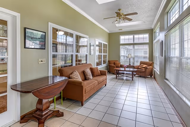 Sunlit common sitting room with brown sofas and armchairs, tiled floor, large windows, and a ceiling fan.