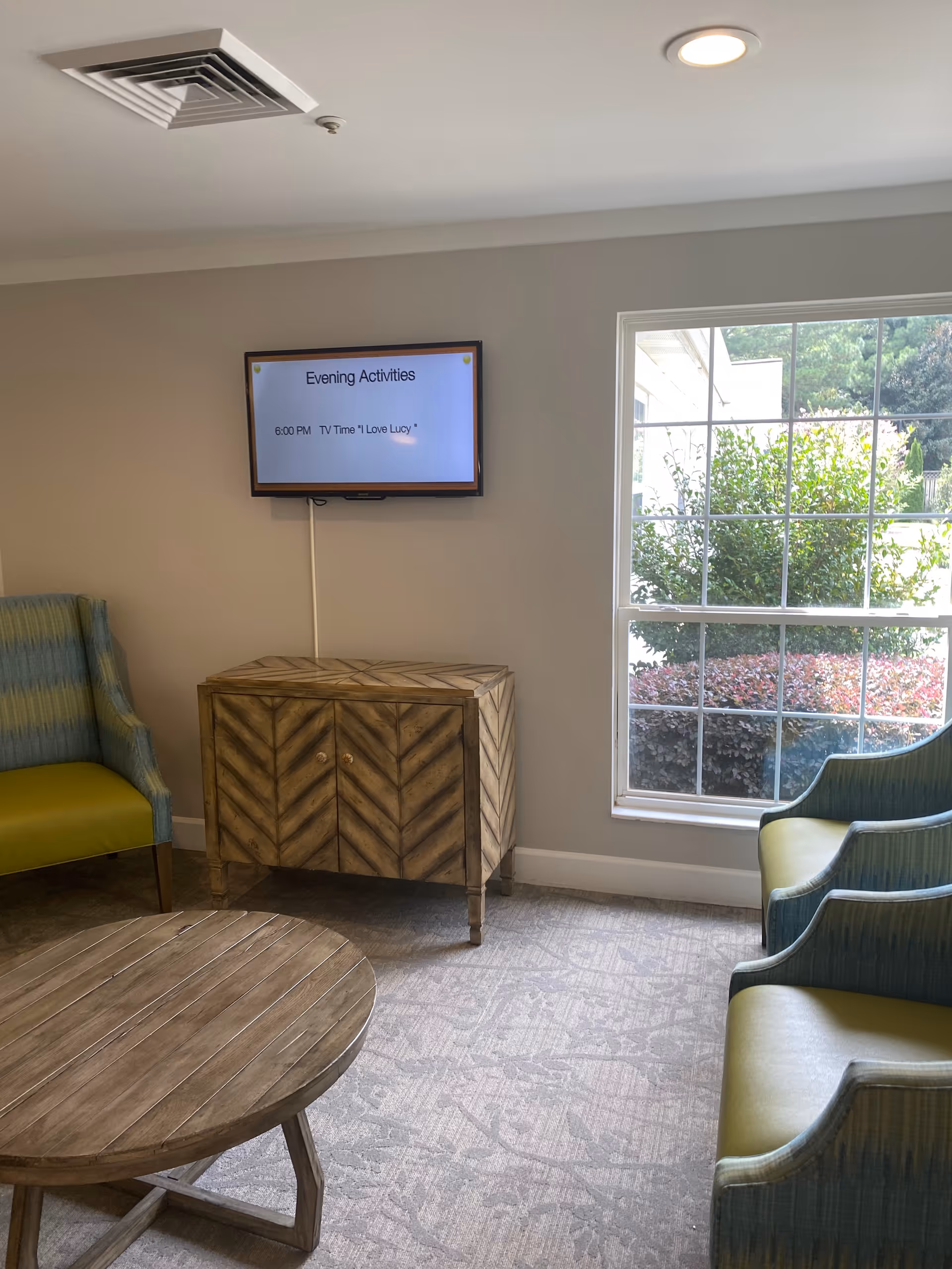 A cozy living room area with a round wooden coffee table in the foreground, two green cushioned chairs on the right, and a green and blue cushioned armchair on the left. A wooden cabinet with a chevron pattern stands against the wall beneath a mounted TV screen displaying 'Evening Activities 6:00 PM TV Time "I Love Lucy"'. A large window on the right lets in natural light and shows greenery outside.