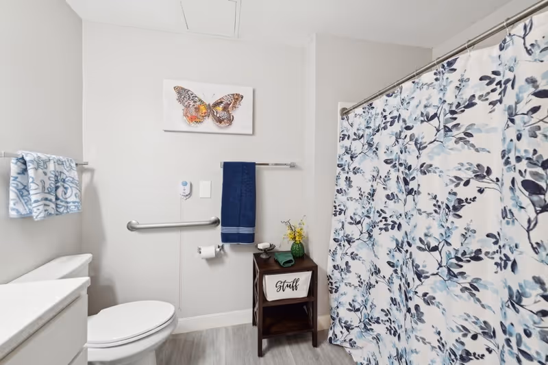 A clean and bright bathroom featuring a white toilet, a countertop with a sink, a towel rack with two towels, a small wooden shelf labeled 'Stuff' holding a green towel and a small vase with yellow flowers, a wall-mounted grab bar, a butterfly artwork on the wall, and a shower with a floral-patterned curtain.