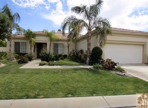 Single-story stucco house with a two-car garage, palm trees and a manicured front lawn under a partly cloudy sky.
