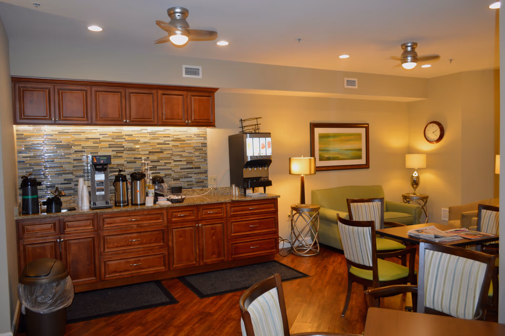 Interior view of a common area in a senior living facility featuring a coffee and beverage station with wooden cabinets and a tiled backsplash. The room includes a green sofa, several chairs with striped upholstery, tables with magazines, two side tables with lamps, a wall clock, and a framed landscape painting. The ceiling has recessed lighting and two ceiling fans.