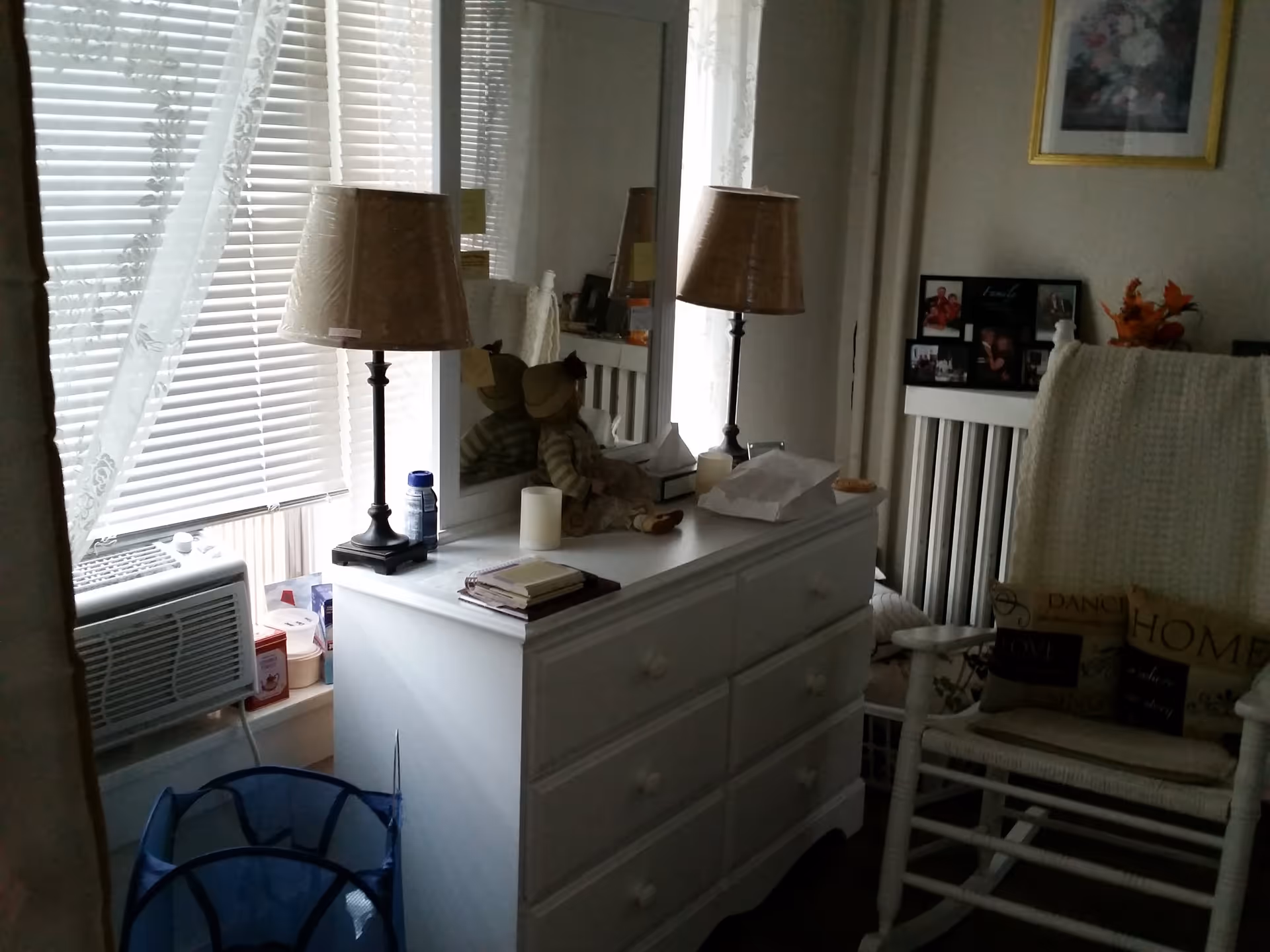A cozy room corner featuring a white dresser with a table lamp, a doll, and various small items on top. Next to the dresser is a window with blinds and sheer curtains, and an air conditioning unit below the window. A white rocking chair with a knitted blanket and decorative pillows is positioned nearby. The wall has framed pictures and a floral artwork.