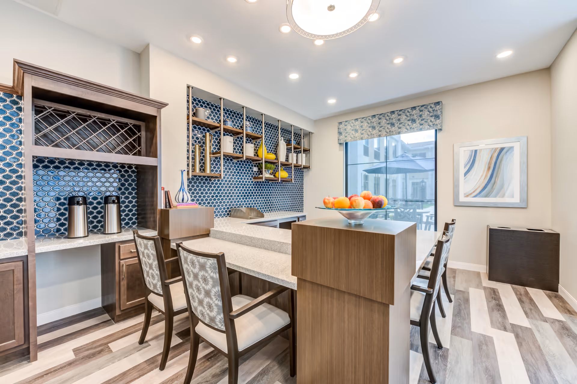 A modern dining area with a wooden table and chairs featuring patterned cushions. The room has a blue tiled backsplash with decorative shelves holding vases and other items. There is a large window with a floral valance, a framed abstract painting on the wall, and a bowl of fruit on the table. The floor has a wood-like pattern, and the ceiling has recessed lighting and a decorative light fixture.