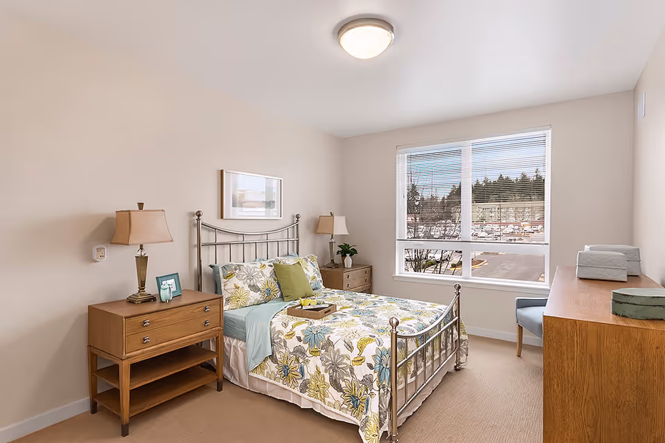 A bright bedroom with a metal frame bed featuring floral bedding in shades of green, blue, and yellow. There are two wooden nightstands with lamps on either side of the bed. A wooden dresser with decorative boxes and a chair are positioned near a large window with blinds, showing a view of trees and parked cars outside. The walls are painted light beige and the room has a beige carpet.