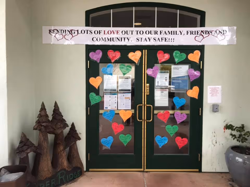 Double green doors decorated with colorful paper hearts and various notices. Above the doors is a banner that reads 'SENDING LOTS OF LOVE OUT TO OUR FAMILY, FRIENDS AND COMMUNITY. STAY SAFE!!!'. To the left of the doors are wooden tree sculptures and a sign that says 'Timber Ridge'. To the right is a large planter with a plant.