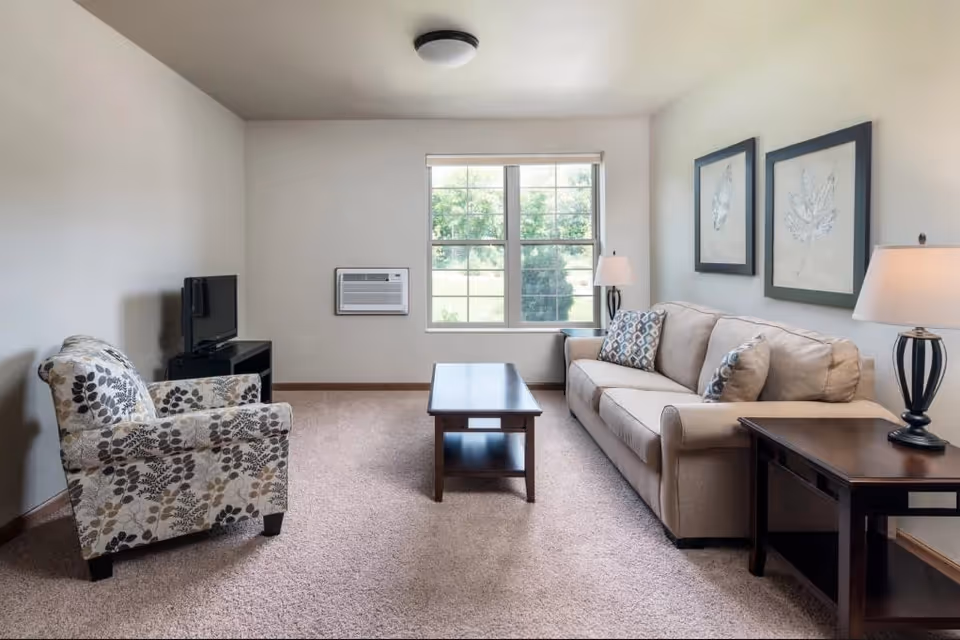 A cozy living room with a beige sofa adorned with patterned throw pillows, a floral-patterned armchair, a wooden coffee table, and two side tables each with a lamp. There are two framed leaf artworks on the wall above the sofa, a window with a view of greenery outside, and a small TV on a stand in the corner.