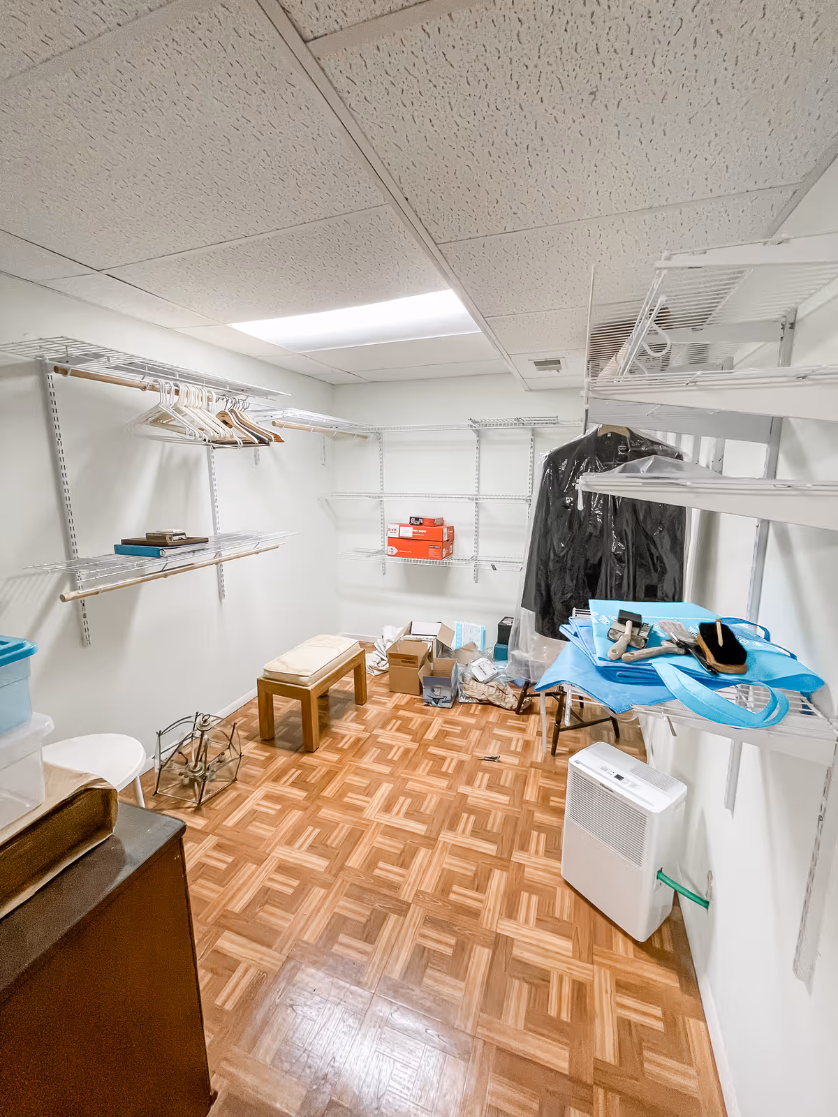 Small storage/closet-like room with wire shelving, hanging garments, boxes, and a dehumidifier on a parquet floor.