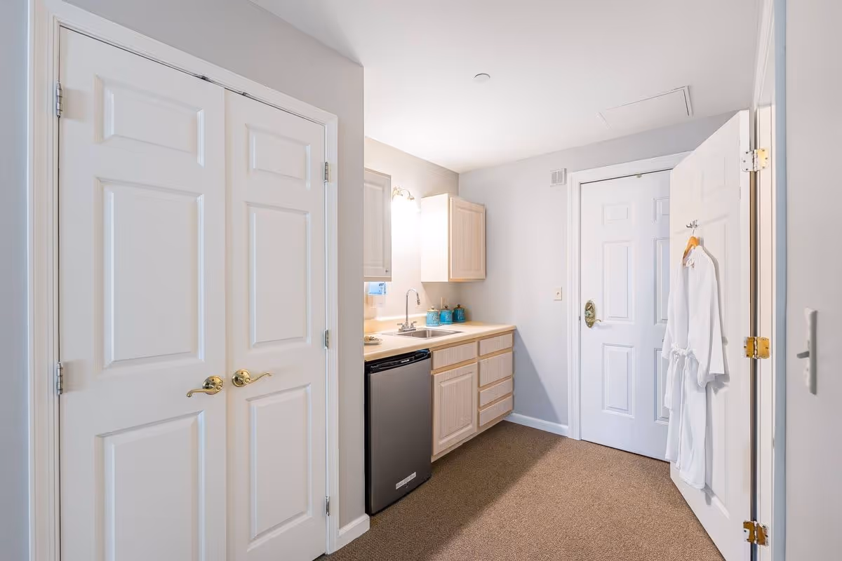 A small kitchenette area with light wood cabinets, a stainless steel mini refrigerator, a sink, and a mirror above the sink. There are two closed white doors on the left and one white door on the right with a white robe hanging on a hook. The walls are painted light gray and the floor is carpeted in a beige color.