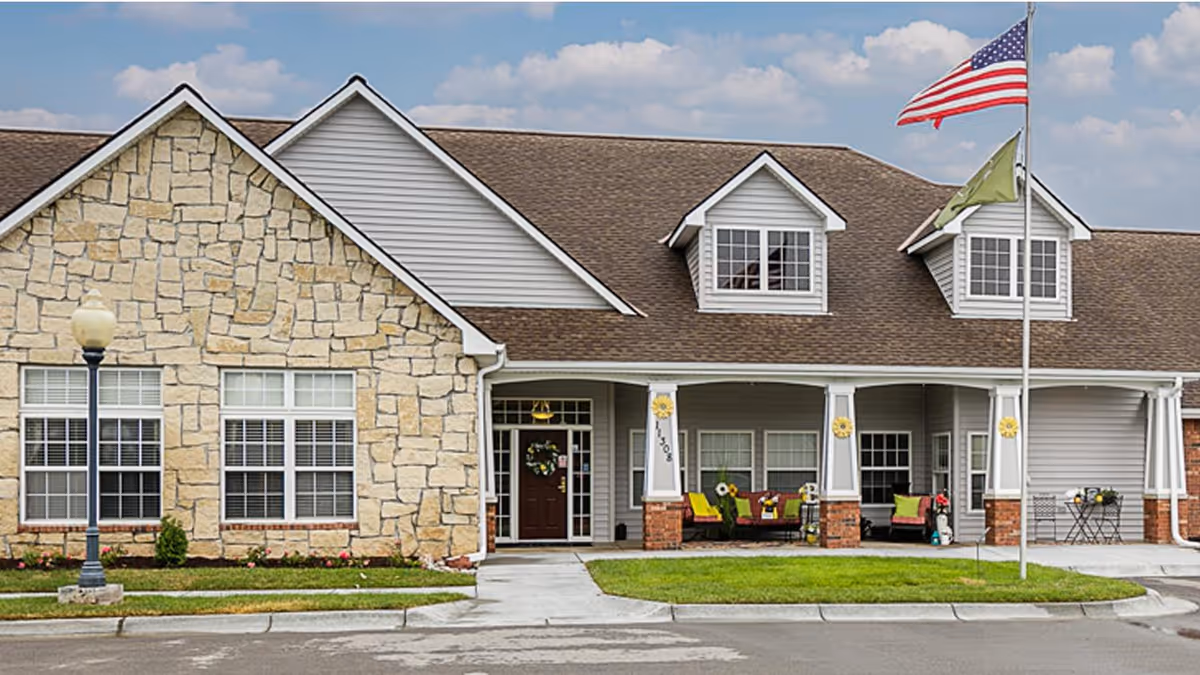 Front exterior view of a single-story senior living facility building with a stone and siding facade, a covered porch with seating, an American flag and another flag on flagpoles, and a well-maintained lawn and sidewalk.