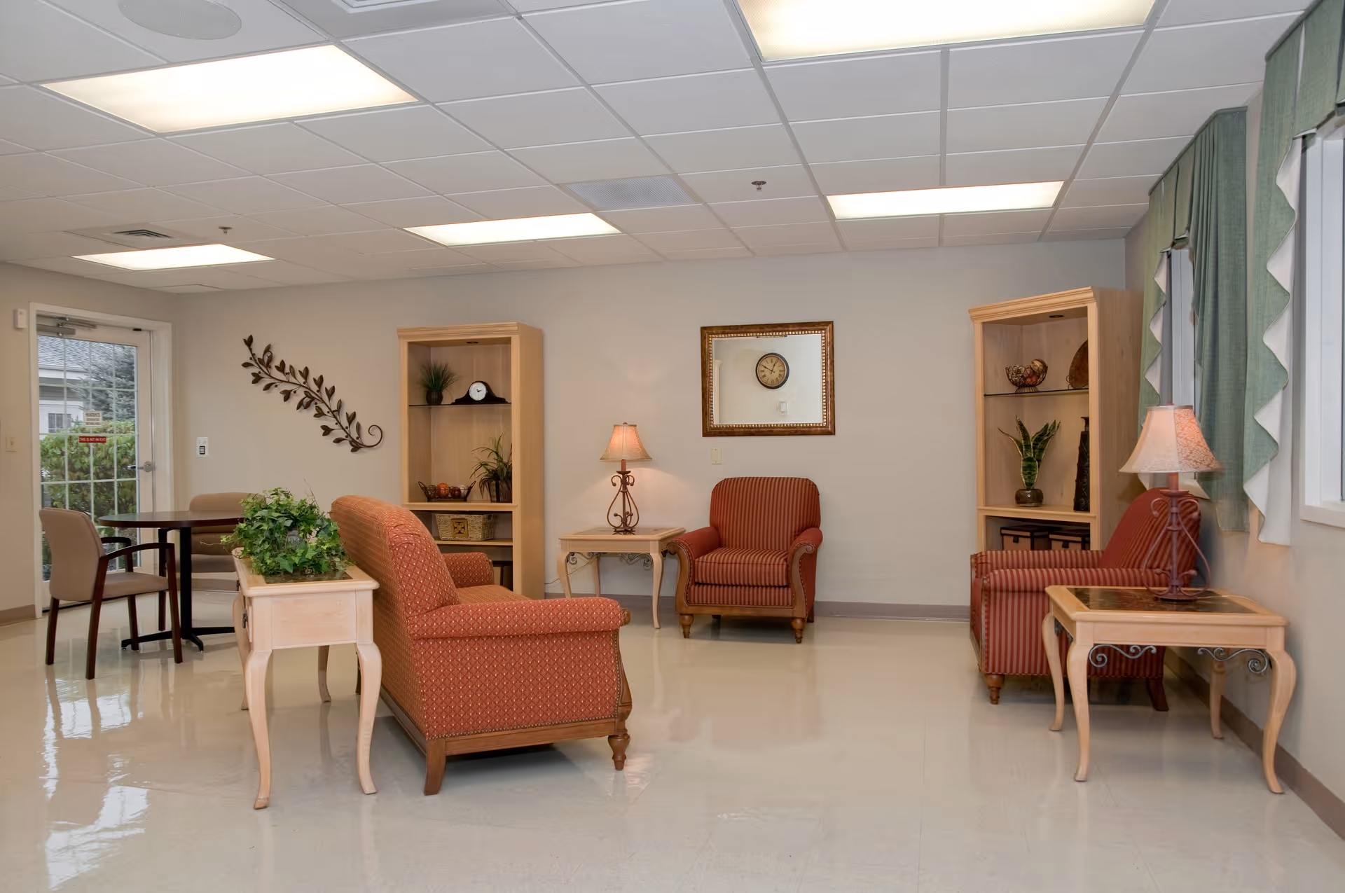 Bright communal sitting room with upholstered armchairs, side tables, lamps, shelving and a small dining table.