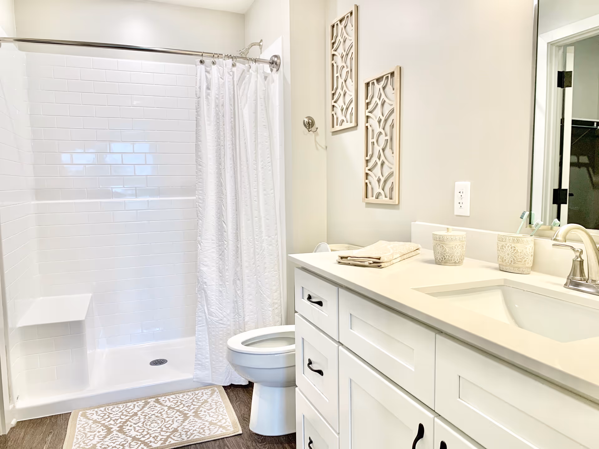 Bright and clean bathroom with a white tiled shower area with a white curtain, a white toilet, a white vanity with a sink, two decorative cups holding toothbrushes, folded towels on the counter, and two decorative wall hangings above the toilet.