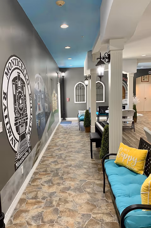 Interior hallway of Arbor Rose of Monticello featuring a mural on the left wall with a Monticello Railway logo and train images. The floor has a stone tile pattern, and the ceiling is painted blue with recessed lighting. On the right side, there are white columns with black lantern-style wall lights, a piano, and seating with turquoise cushions and yellow pillows, one pillow reads 'my happy place'.