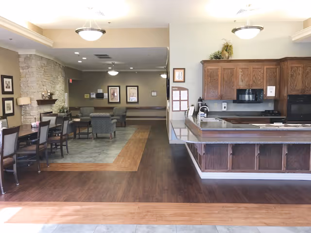 Interior view of a senior living facility showing a dining area with tables and chairs on the left, a lounge area with armchairs in the background, and a kitchen area with wooden cabinets and a countertop on the right. The floor has a combination of wood and tile, and the walls are decorated with framed pictures and a stone accent wall.