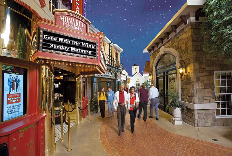 A group of elderly people walking along a street inside a senior living facility designed to look like an outdoor town square with a Monarch Cinema marquee advertising 'Gone With the Wind Sunday Matinee'. The area is well-lit with storefronts, a clock tower in the background, and a starry sky ceiling.