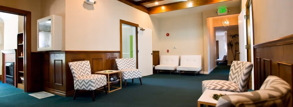 A spacious interior common area with green carpet, wood-paneled walls, and a ceiling with recessed lighting. The room features several chevron-patterned armchairs arranged around small tables, and two white cushioned chairs against the far wall. A hallway with an exit sign is visible in the background.