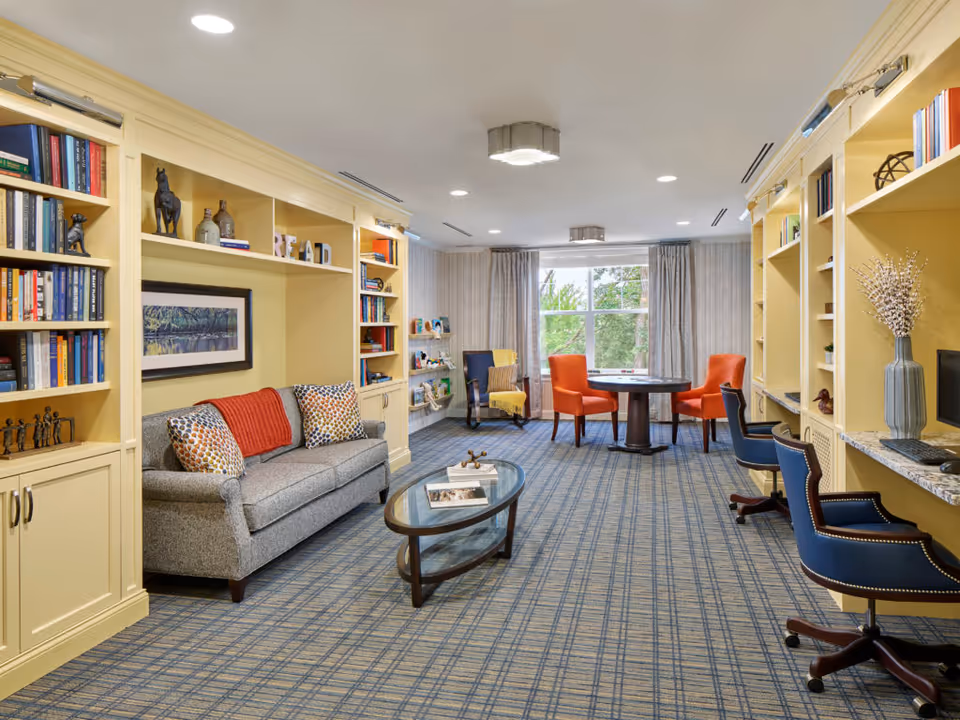 A cozy and well-lit common area featuring built-in yellow bookshelves filled with books and decorative items. A gray sofa with patterned pillows and a red throw blanket sits against one wall. In the center, there is a glass coffee table with books and decorative pieces. Toward the back, a round table with four chairs, two orange and two blue, is positioned near a large window with gray curtains, allowing natural light to brighten the room. The carpet has a blue and beige checkered pattern, and the ceiling has recessed lighting and two modern light fixtures.