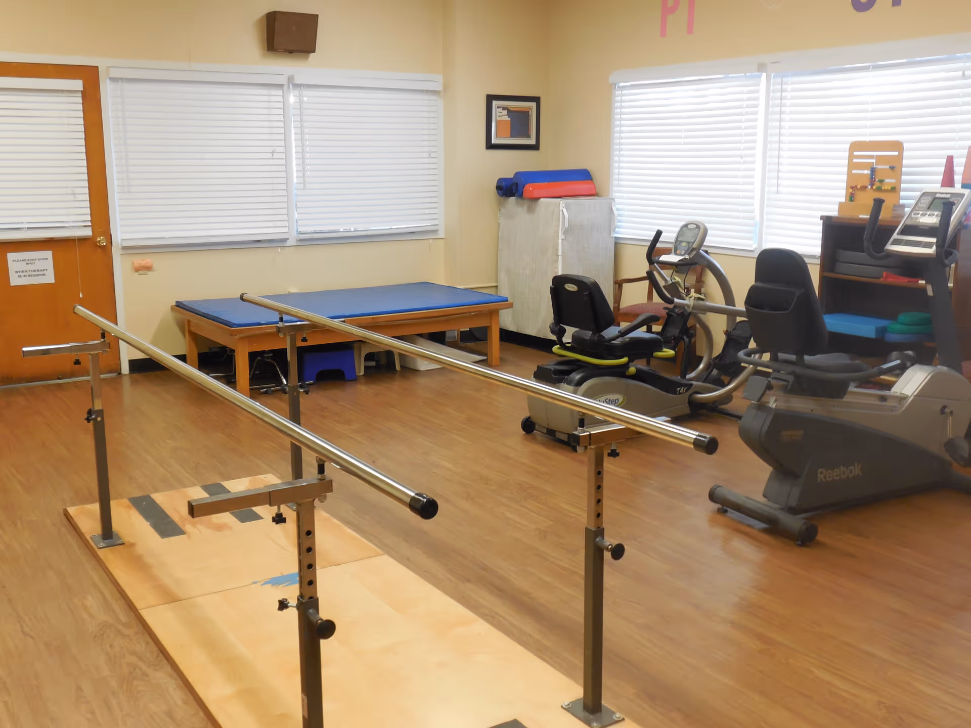 Physical therapy room with parallel bars for walking exercises, a padded therapy table, exercise bikes, and various therapy equipment against the walls. The room has wooden flooring and windows with white blinds.