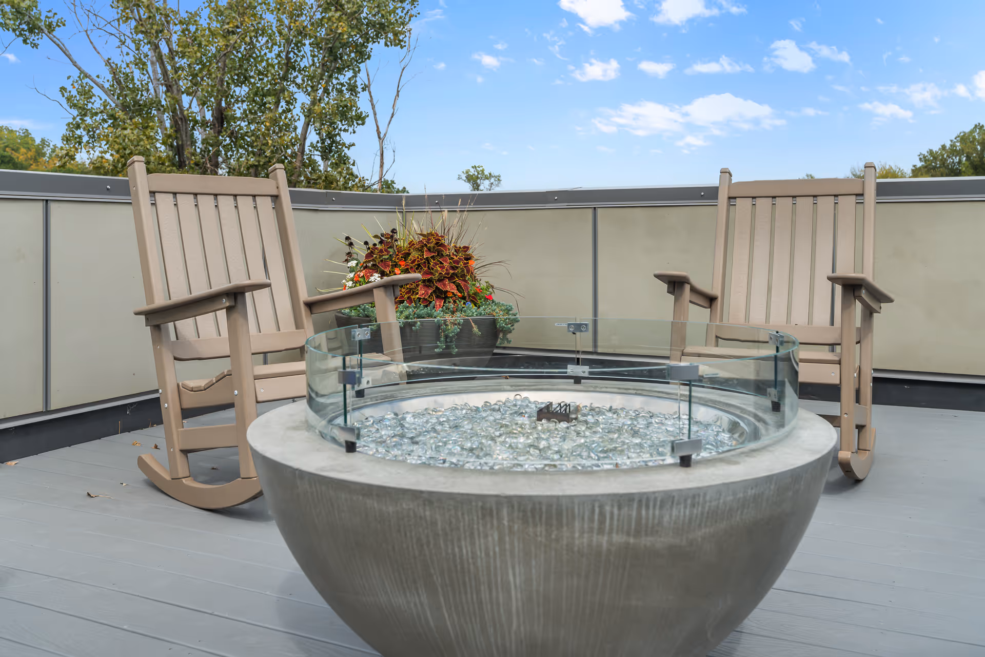 Rooftop patio with two wooden rocking chairs, a round concrete fire pit with a glass surround, and a planter of flowers under a blue sky.