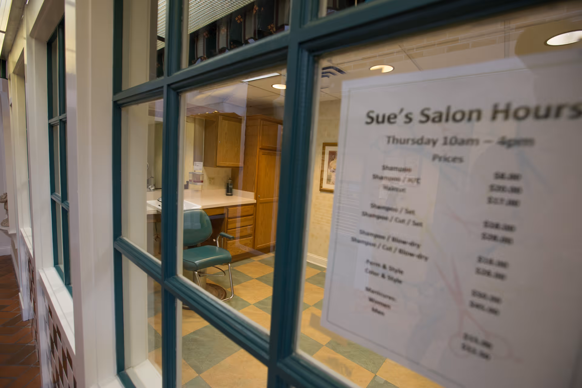 View through a window into a salon room with a green salon chair, wooden cabinets, and a countertop. A sign on the window displays Sue's Salon Hours and price list for various hair services.