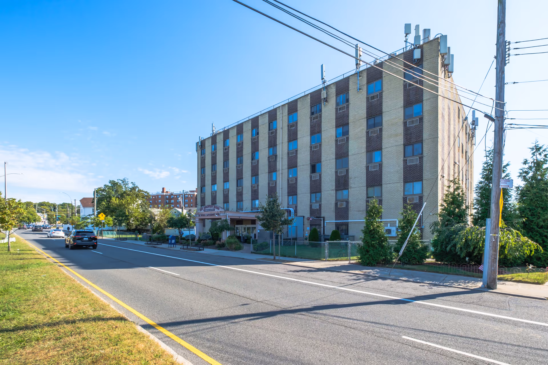 Exterior view of a multi-story brick building with several windows and air conditioning units, situated along a street with cars driving by and trees lining the sidewalk under a clear blue sky.
