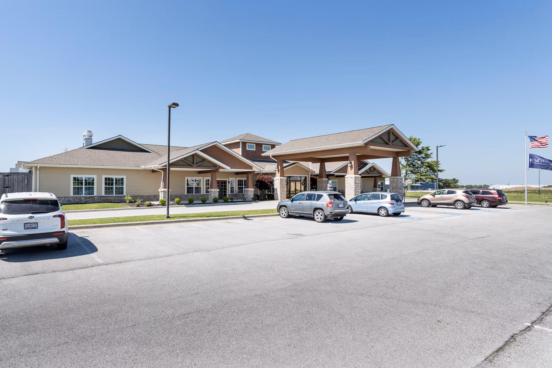 Exterior view of Oak Pointe of Carthage senior living facility showing a single-story building with a covered entrance, several parked cars in the parking lot, a clear blue sky, and two flags on flagpoles to the right.