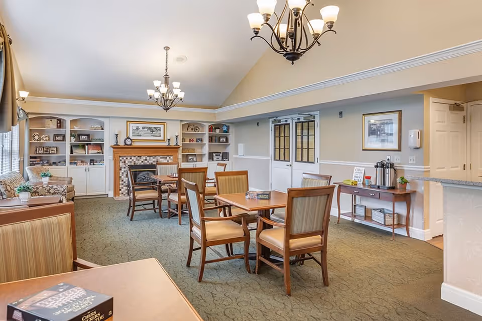 A well-lit common area in a senior living facility featuring multiple wooden tables with chairs, a fireplace with a tiled surround, built-in shelves with decorative items, and a side table with a coffee station. The room has beige walls, carpeted floor, and two chandeliers hanging from the ceiling.