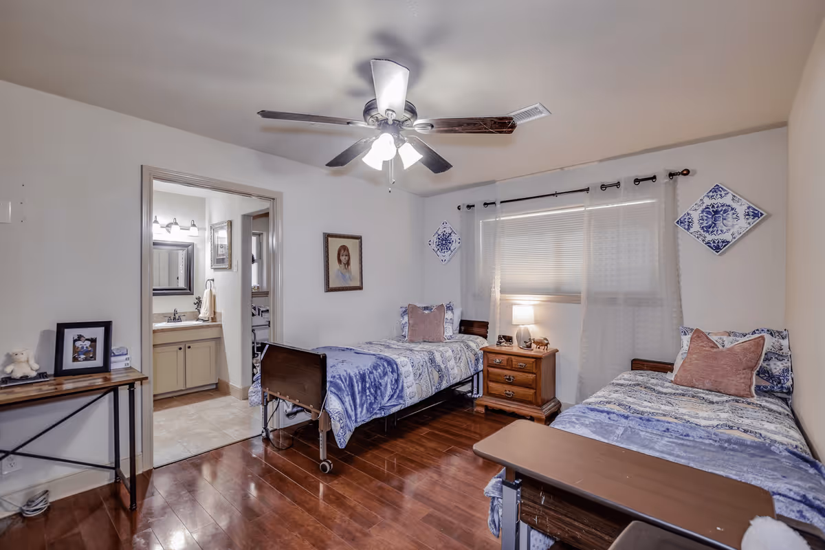 A bedroom with two single beds, each with blue and white bedding and a pink pillow. There is a wooden nightstand with a lamp between the beds. A ceiling fan with lights is on the ceiling. A window with sheer curtains is behind the nightstand. A small table with framed photos and a stuffed animal is on the left side of the room. An open door reveals a bathroom with a sink and mirror.