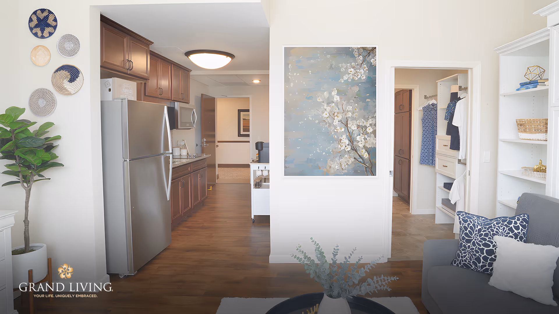 Open living room with a gray sofa, decorative pillows and artwork, looking into a kitchen with stainless refrigerator and a walk-in closet.