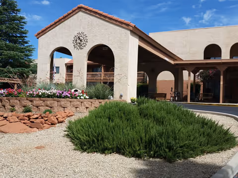 Exterior view of Sedona Winds Assisted Living Community featuring a beige stucco building with a red tile roof, arched entryways, landscaped garden with green shrubs, colorful flowers, and a clear blue sky.