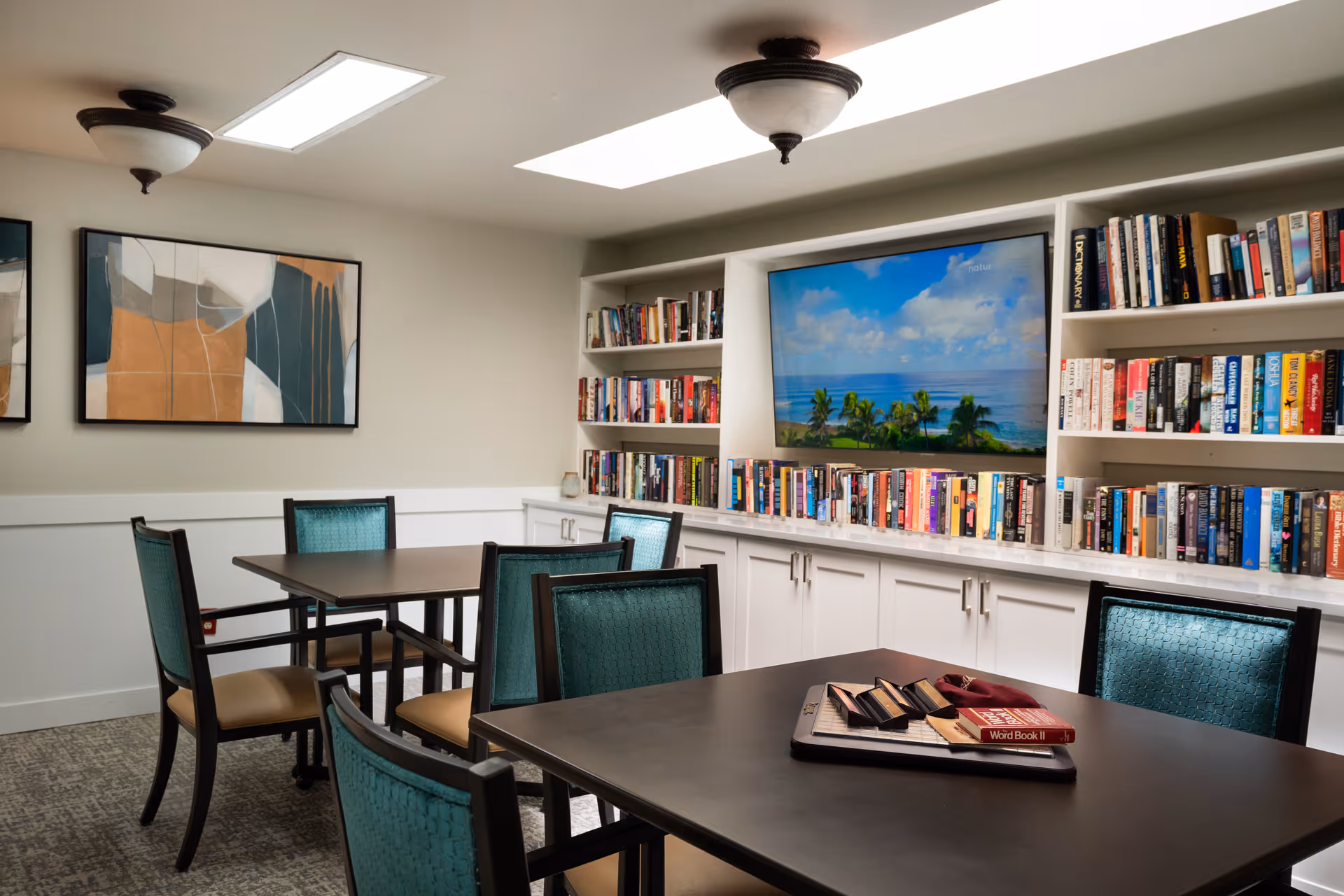 A cozy reading or activity room with several tables and teal cushioned chairs. The room features built-in white bookshelves filled with books and a large flat-screen TV displaying a scenic beach view. Abstract artwork is hung on the wall, and the ceiling has two light fixtures and a skylight.