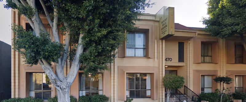 Exterior view of Beverly Hills Gardens senior living facility showing a two-story building with beige walls, multiple windows with curtains, a sign displaying the facility name, and trees and shrubs in front.
