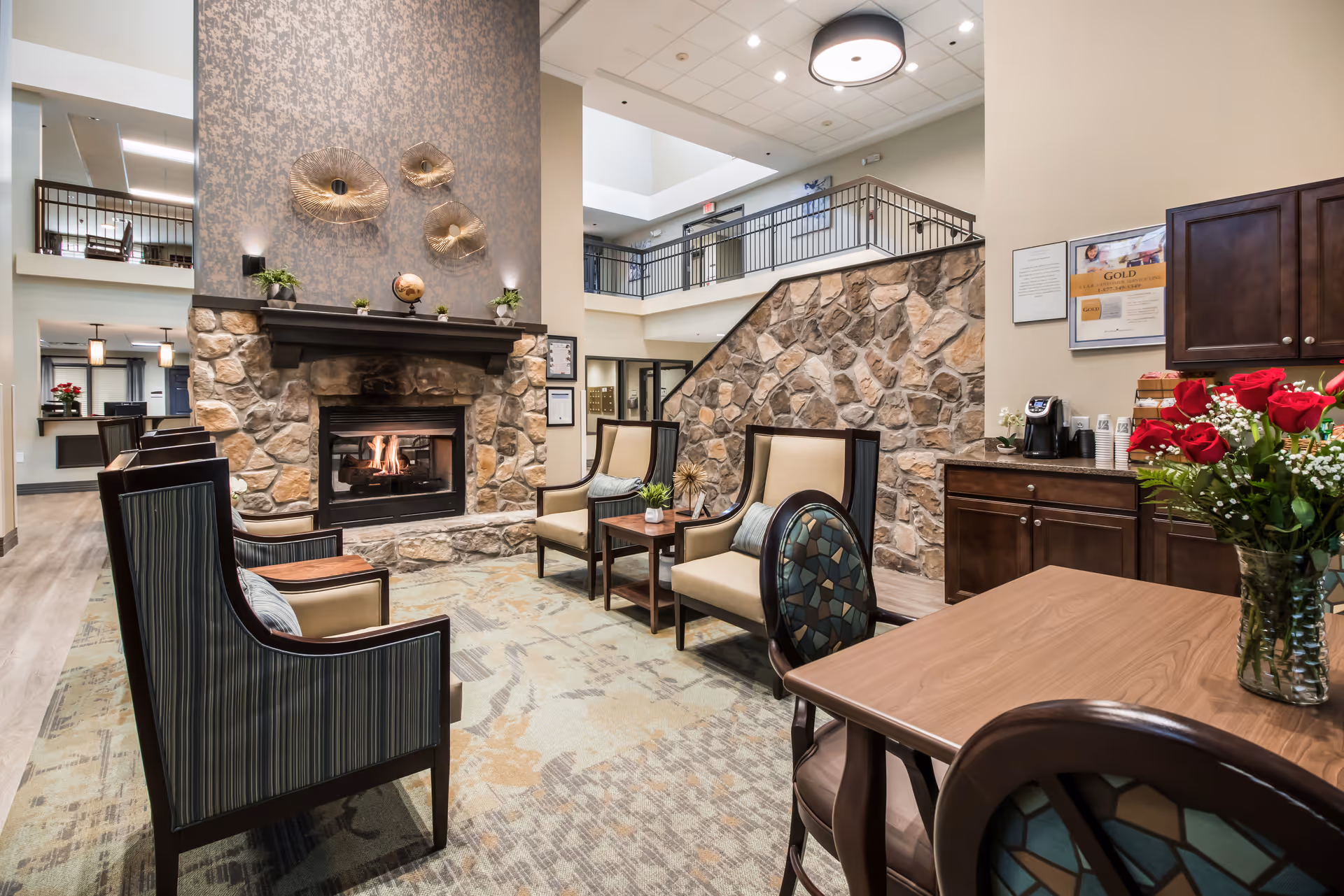 A cozy seating area in a senior living facility featuring a stone fireplace with a fire burning, surrounded by four armchairs and a small table. The room has a high ceiling with a balcony railing visible on the upper level. There is a wooden table with a vase of red roses in the foreground and a coffee station on a cabinet against the wall.
