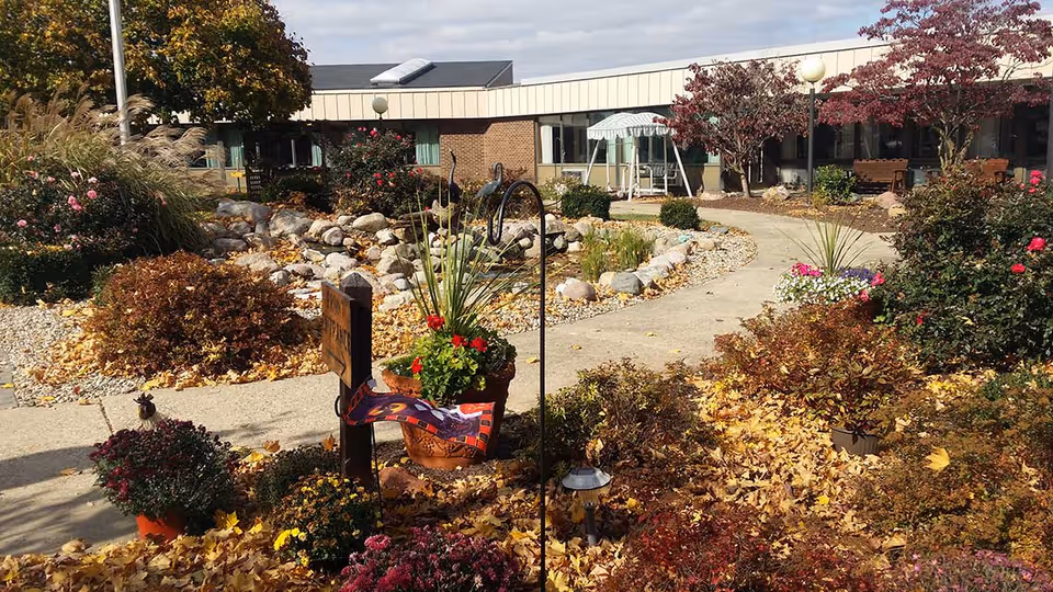 Outdoor garden area at Pleasant Meadows Senior Living with a paved walkway, various plants and flowers, a small rock garden, and a building in the background under a partly cloudy sky.