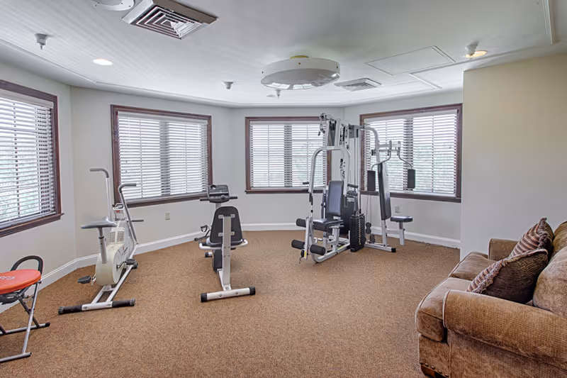 A bright assisted-living fitness room with stationary bikes, a multi-station weight machine, and a sofa by bay windows.